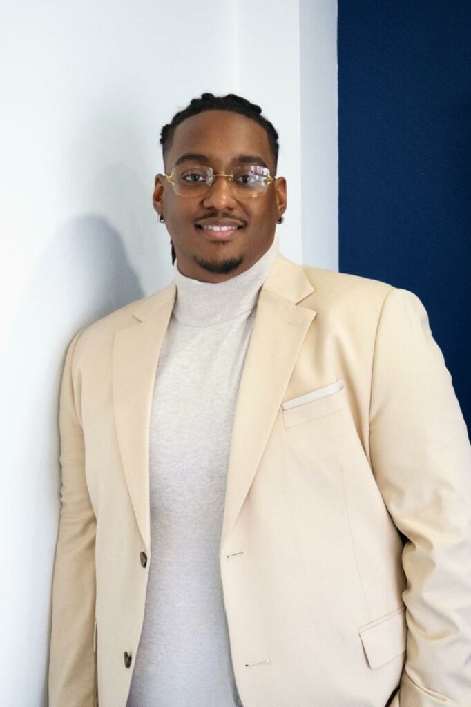 Carlito Henry vêtu de beige, regard direct, mur bleu en arrière-plan